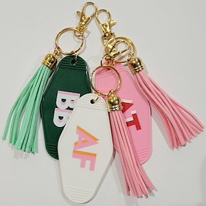 May include: Three keychains with different coloured motel key tags. The green key tag says "BB", the pink key tag says "AT", and the white key tag says "AF". Each keychain has a gold key ring and a tassel.