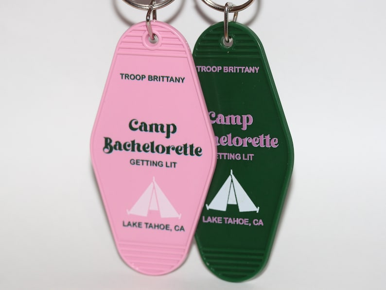 May include: Two pink and green keychains shaped like motel room keys. The keychains say "Camp Bachelorette Getting Lit" and "Camp Bachelorette Getting Lit" with a tent graphic and "Lake Tahoe, CA".