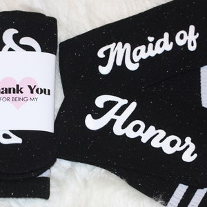 K&ouml;nnte beinhalten: Zwei Paar schwarze Socken mit wei&szlig;en Streifen. Ein Paar sagt "Maid of Honor" und das andere sagt "&" mit einem Dankes-Etikett, auf dem steht "Danke, dass du meine bist".