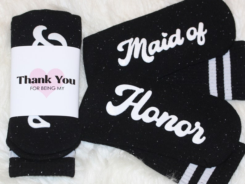 K&ouml;nnte beinhalten: Zwei Paar schwarze Socken mit wei&szlig;en Streifen. Ein Paar sagt "Maid of Honor" und das andere sagt "&" mit einem Dankes-Etikett, auf dem steht "Danke, dass du meine bist".