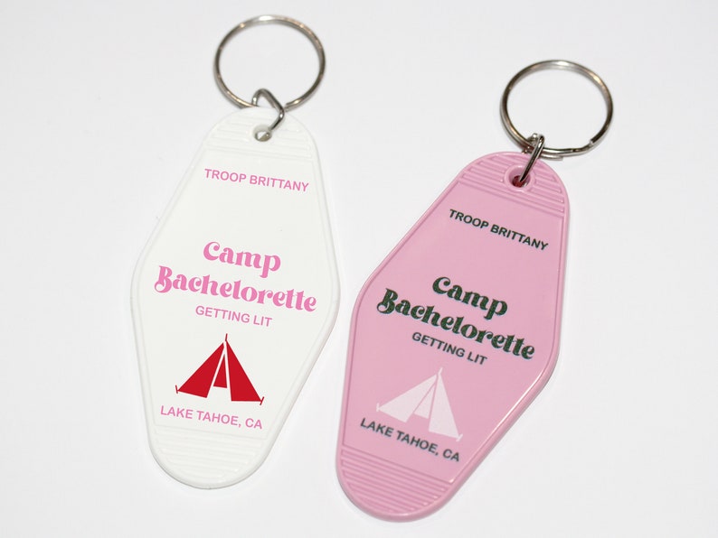 May include: Two keychains shaped like motel room keys. One is white with pink text and a red tent graphic. The other is pink with green text and a white tent graphic. Both say "Camp Bachelorette Getting Lit" and "Lake Tahoe, CA".