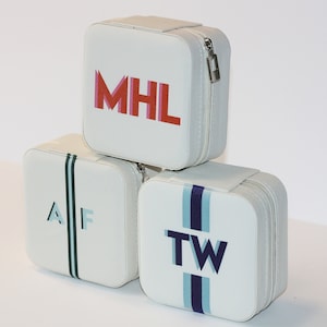 May include: Three white square-shaped zippered travel cases with different colored initials. The top case has the initials "MHL" in red, pink, and orange. The middle case has the initials "AF" in light blue and dark green. The bottom case has the initials "TW" in light blue and dark purple.