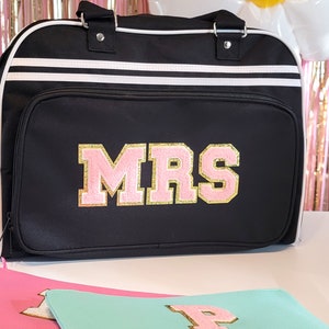 May include: Black duffel bag with white stripes and the word "MRS" in pink and gold lettering. The bag has a top handle and a front pocket. Additional matching pouches are visible in the foreground.