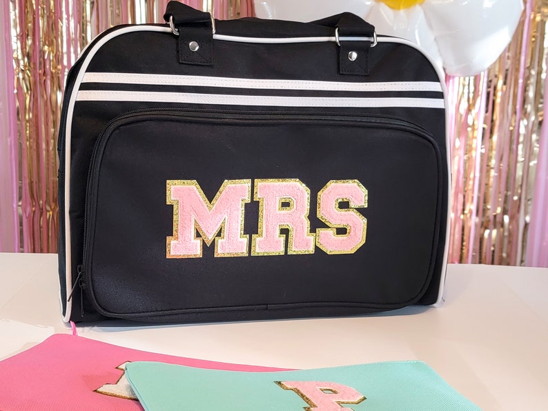 May include: Black duffel bag with white stripes and the word "MRS" in pink and gold lettering. The bag has a top handle and a front pocket. Additional matching pouches are visible in the foreground.