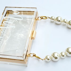 May include: A clear acrylic clutch with gold-tone trim and hardware. The clutch features a pearl beaded handle and the name "Mrs. Broadbent" in white script. The rectangular box-shaped purse has a clasp closure.