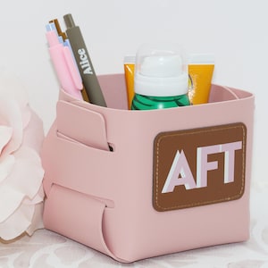 May include: A pink desk organizer with a brown leather patch that reads "AFT" in white letters. The organizer holds pens, a small bottle, and tubes of lotion. The pens are pink and gray, and one is labeled "Alice".