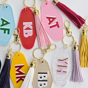 May include: A selection of keyrings in various colours, each with gold-coloured fixings and tassels. The keyrings are in shades of blue, red, pink, yellow, black, and beige. Each keyring displays a different set of initials in a contrasting colour.