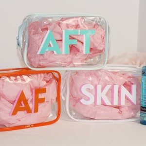 May include: Three clear plastic zippered pouches with pink tissue paper inside. The pouches are labeled "AFT", "AF", and "SKIN" in colorful letters. Two bottles of skincare products are also shown.