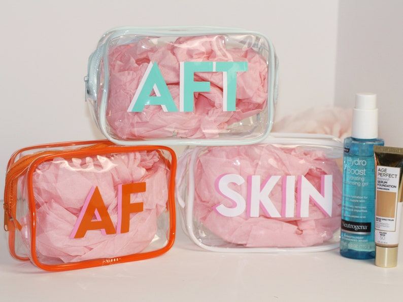 May include: Three clear plastic zippered pouches with pink tissue paper inside. The pouches are labeled "AFT", "AF", and "SKIN" in colorful letters. Two bottles of skincare products are also shown.