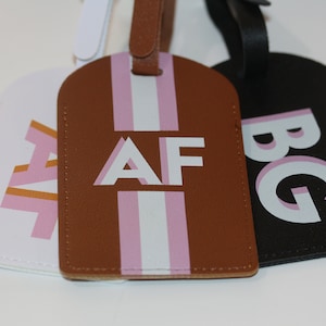 May include: Three rectangular luggage tags with rounded tops and matching straps. The white tag features pink and orange letters AF, the brown tag has white and pink stripes with white letters AF, and the black tag displays white and pink letters BG.