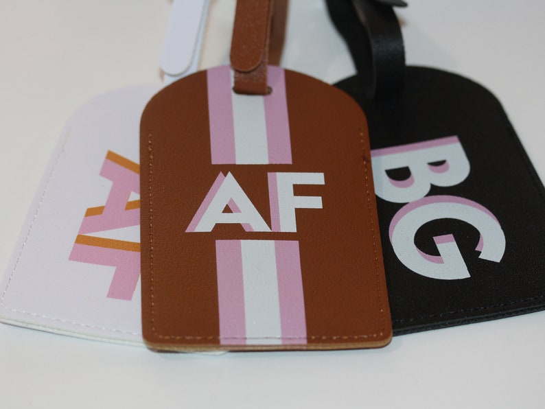May include: Three rectangular luggage tags with rounded tops and matching straps. The white tag features pink and orange letters AF, the brown tag has white and pink stripes with white letters AF, and the black tag displays white and pink letters BG.