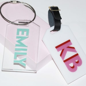 May include: Two clear acrylic luggage tags with black straps. One tag has the name "EMILY" in teal lettering. The other tag has the initials "KB" in pink and red lettering.