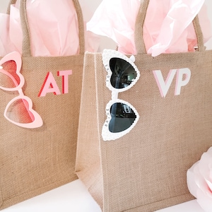 May include: Two burlap tote bags with pink tissue paper inside. One bag has pink heart-shaped sunglasses and the letters "AT" in pink and red. The other bag has white heart-shaped sunglasses and the letters "VP" in pink and white.