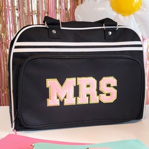 May include: Black duffel bag with white stripes and trim. The bag features the word "MRS" in pink and gold varsity letters. The bag has a top handle and a front pocket.