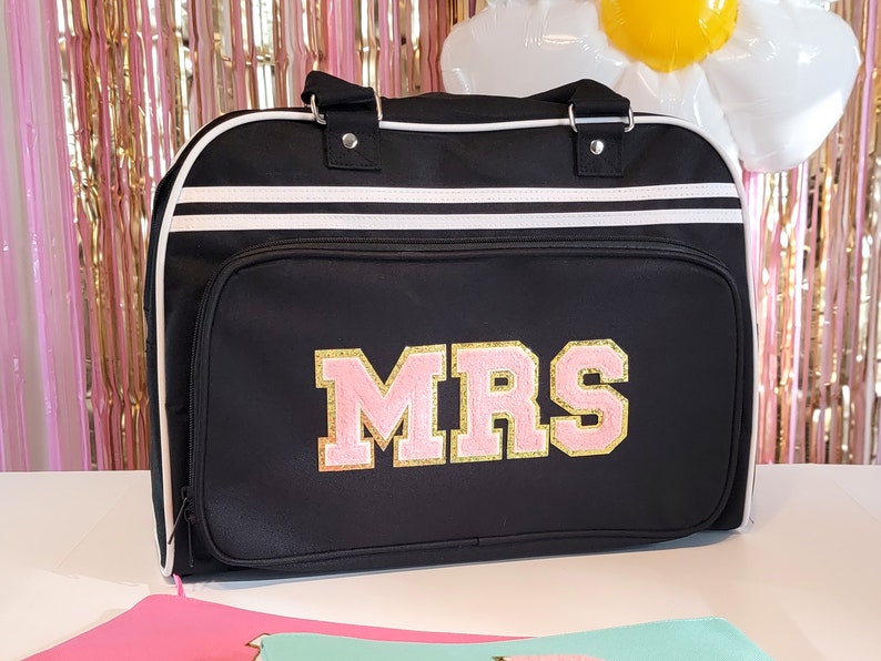 May include: Black duffel bag with white stripes and trim. The bag features the word "MRS" in pink and gold varsity letters. The bag has a top handle and a front pocket.