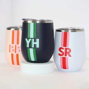 May include: Three stainless steel wine tumblers with personalized initials and colorful stripes. The tumblers are in navy blue, white, and pink.