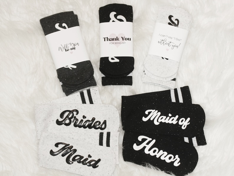 K&ouml;nnte beinhalten: Set von vier Paar Socken mit verschiedenen Botschaften. Zwei Paar sind schwarz mit wei&szlig;em Text und zwei Paar sind wei&szlig; mit schwarzem Text. Die Botschaften auf den Socken sind "Will you be my", "Thank you for being my", "I can't say 'I Do' without you", "Brides Maid", "Maid of Honor".