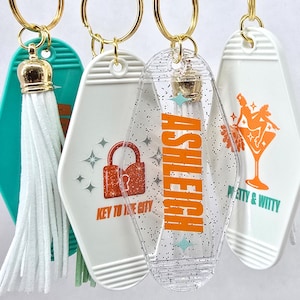 May include: Four keychains with gold rings. One is teal with a gold crown and white tassel. The others are white and clear with various designs and text, including a lock, the name "ASHLEIGH", and a cocktail glass.