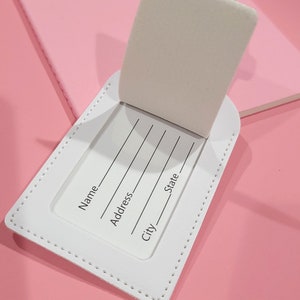 May include: A white rectangular luggage tag with a flip-up cover revealing an identification card. The card features printed lines and text labels for Name, Address, City, and State. The tag has visible white stitching along the edges.