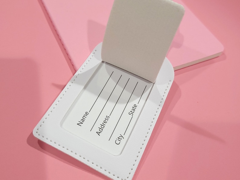 May include: A white rectangular luggage tag with a flip-up cover revealing an identification card. The card features printed lines and text labels for Name, Address, City, and State. The tag has visible white stitching along the edges.