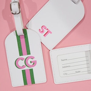 May include: Three white luggage tags with silver buckles. One tag features a vertical green and pink stripe with the letters CG. Another tag displays the letters ST in pink. The third tag shows a clear window with lines for Name, Address, City, and State.