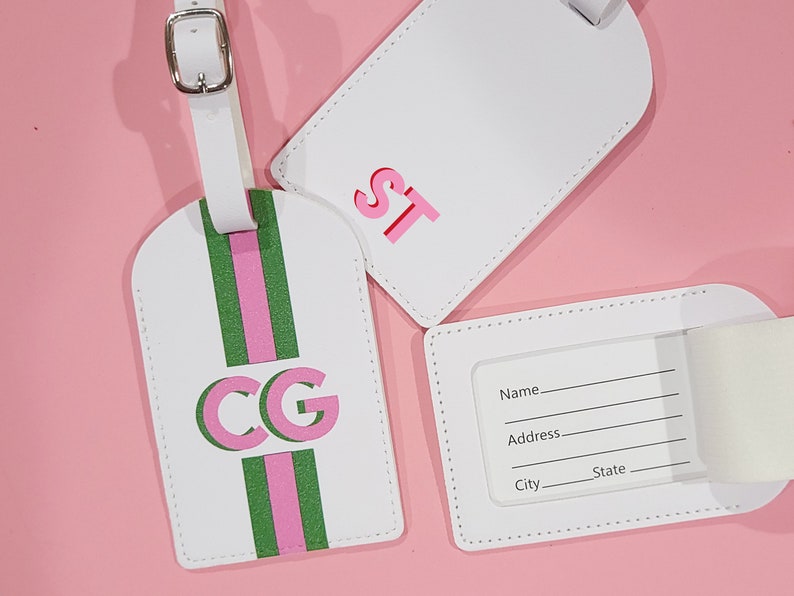 May include: Three white luggage tags with silver buckles. One tag features a vertical green and pink stripe with the letters CG. Another tag displays the letters ST in pink. The third tag shows a clear window with lines for Name, Address, City, and State.
