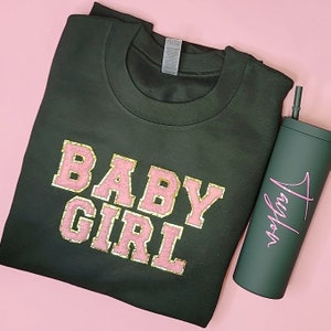 May include: A dark green sweatshirt with the words "BABY GIRL" in pink letters with gold glitter accents. The sweatshirt is folded and a dark green tumbler with the word "Taylor" in pink script is next to it.