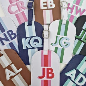 May include: A collection of rectangular luggage tags in various colors including white, navy, black, and brown. Each tag features a vertical stripe, a silver-toned buckle strap, and two-letter initials printed in bold, layered block lettering.