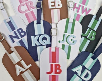 May include: A collection of rectangular luggage tags in various colours including white, navy, black, and brown. Each tag features a vertical stripe, a silver-toned buckle strap, and two-letter initials printed in bold, layered block lettering.