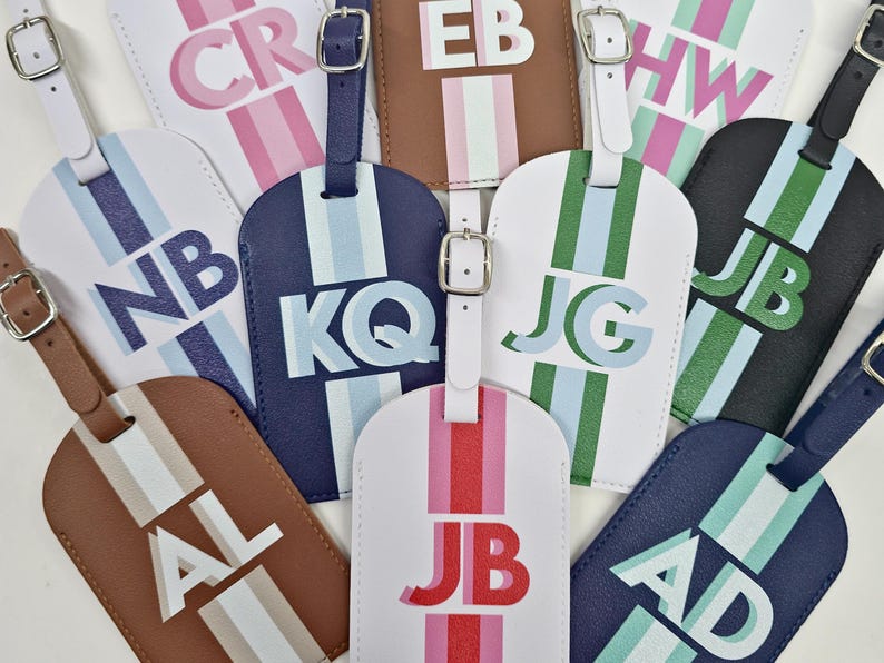 May include: A collection of rectangular luggage tags in various colors including white, navy, black, and brown. Each tag features a vertical stripe, a silver-toned buckle strap, and two-letter initials printed in bold, layered block lettering.
