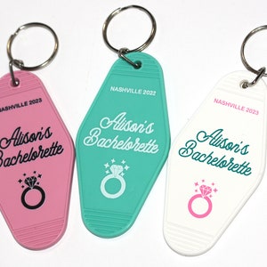 May include: Three keychains with a motel key design. Each keychain is a different color: pink, teal, and white. The keychains have the text "Alison's Bachelorette" and "Nashville 2023" printed on them.
