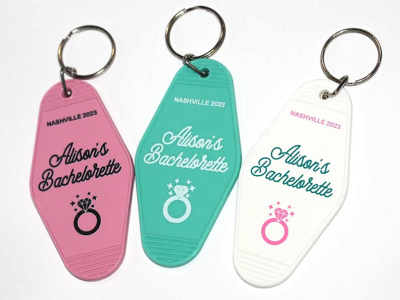 May include: Three keychains with a motel key design. Each keychain is a different color: pink, teal, and white. The keychains have the text "Alison's Bachelorette" and "Nashville 2023" printed on them.