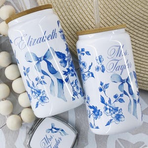 May include: Two white ceramic can-shaped cups with blue floral designs and blue ribbons. The cups have personalized names, "Elizabeth" and "Miss Taylor".