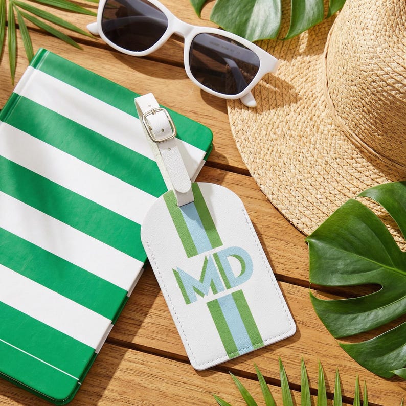May include: A white luggage tag featuring a vertical green and light blue stripe pattern with the monogram MD in the center. The tag includes a white adjustable strap with a silver-toned metal buckle, designed for identifying travel bags.