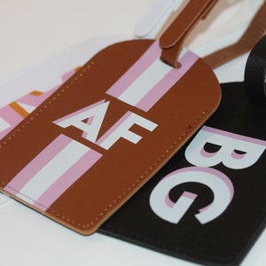 May include: Three rectangular luggage tags with rounded tops and matching straps. One tag is brown with white and pink vertical stripes and the letters AF. One is black with the letters BG. One is white with a pink and orange geometric pattern.