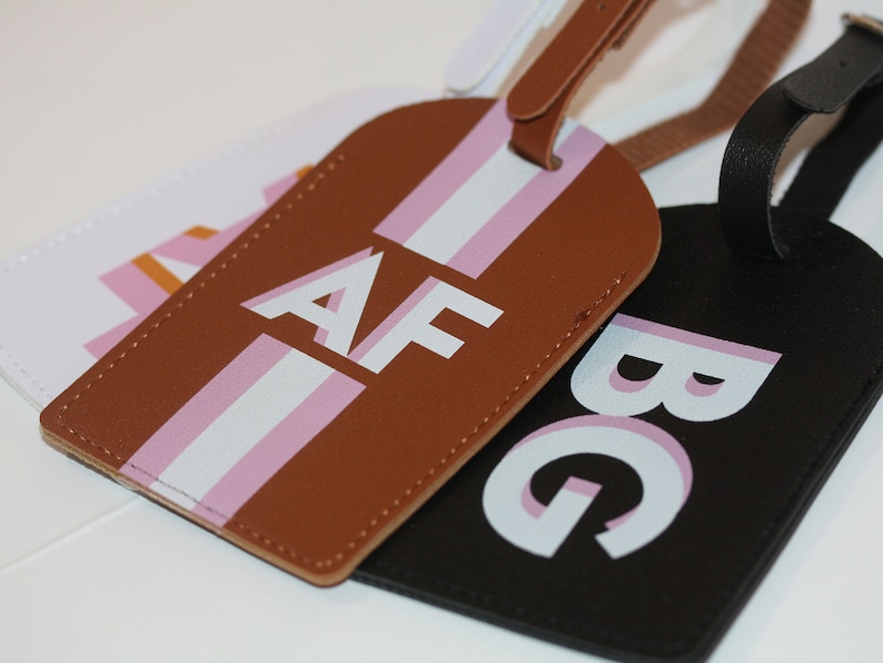 May include: Three rectangular luggage tags with rounded tops and matching straps. One tag is brown with white and pink vertical stripes and the letters AF. One is black with the letters BG. One is white with a pink and orange geometric pattern.