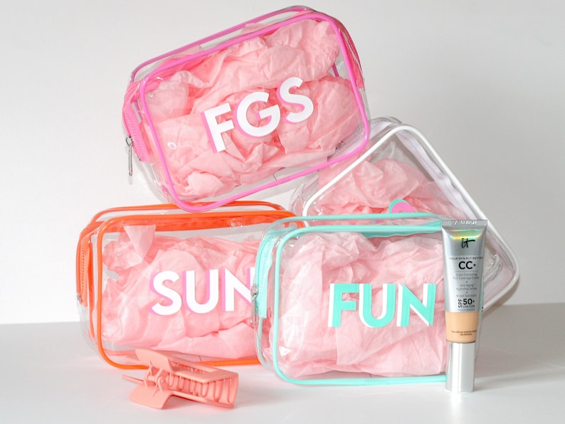 May include: Four clear plastic zippered bags with pink tissue paper inside. The bags have different colored trim and the words "FGS", "SUN", "FUN" printed on them. A tube of IT Cosmetics CC+ Cream with SPF 50 is next to the bag with the word "FUN" printed on it. A pink hair clip is in front of the bag with the word "SUN" printed on it.
