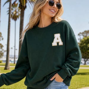 May include: A dark forest green long-sleeve crewneck sweatshirt featuring a large, white, varsity-style letter A patch on the upper left chest. The sweatshirt has a relaxed fit with ribbed cuffs and a ribbed neckline, made of a solid fabric.