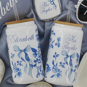 May include: Two white ceramic canisters with blue floral designs and personalized names. The canisters have bamboo lids and clear straws. The canisters are labeled "Elizabeth" and "Mrs. Taylor".