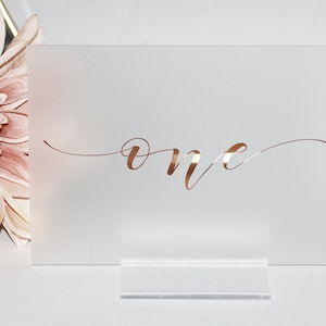 May include: White acrylic table number with a rose gold 'one' written in cursive.