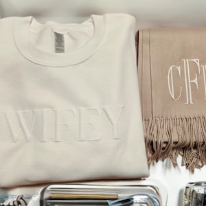 May include: A cream-colored sweatshirt with the word "WIFEY" embroidered on the front, and a brown scarf with the monogram "CFD" embroidered on it.