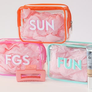 May include: Three clear plastic zippered bags with pink, orange, and turquoise trim. Each bag has a different word printed on it: "SUN", "FGS", and "FUN". A pink hair clip and a tube of foundation are also pictured.