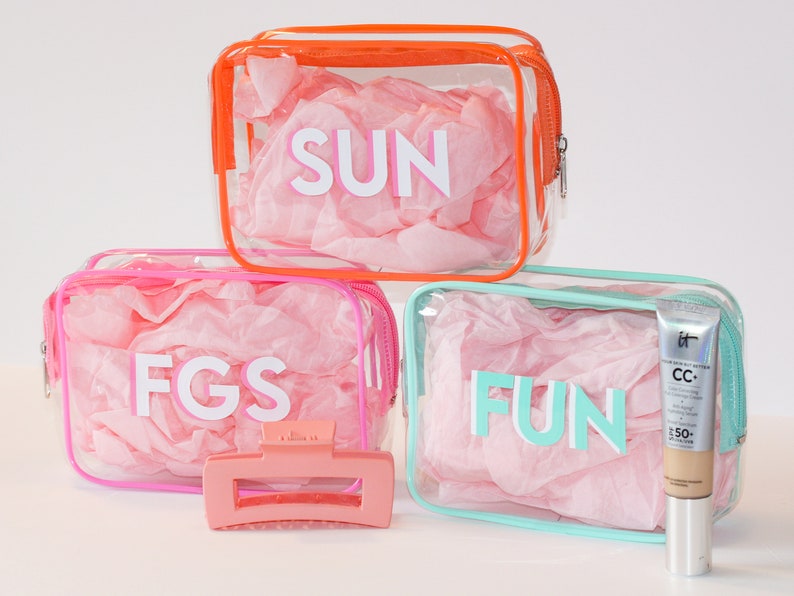 May include: Three clear plastic zippered bags with pink, orange, and turquoise trim. Each bag has a different word printed on it: "SUN", "FGS", and "FUN". A pink hair clip and a tube of foundation are also pictured.