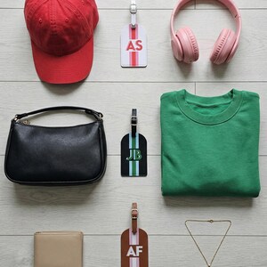 May include: A red baseball cap, a white luggage tag with AS, pink headphones, a black handbag, a black luggage tag with JB, a green sweatshirt, a beige wallet, a brown luggage tag with AF, and a thin gold chain necklace arranged on a light surface.
