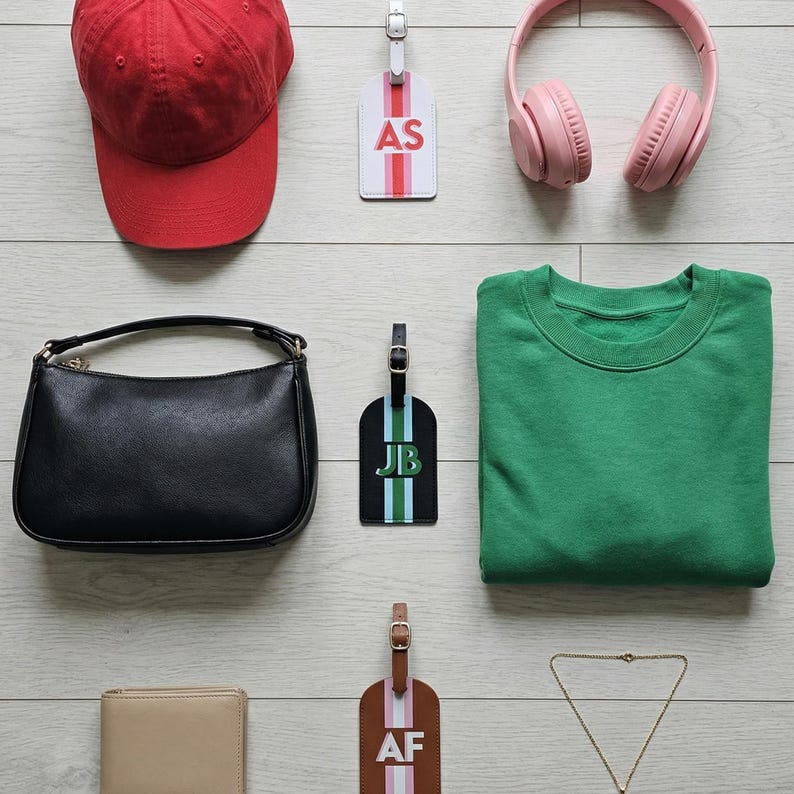 May include: A red baseball cap, a white luggage tag with AS, pink headphones, a black handbag, a black luggage tag with JB, a green sweatshirt, a beige wallet, a brown luggage tag with AF, and a thin gold chain necklace arranged on a light surface.