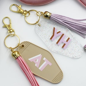 May include: Two keyrings with gold-coloured hardware and colourful tassels. One keyring is clear with glitter and the letters "YH" in brown and pink. The other is tan with the letters "AT" in pink and white.