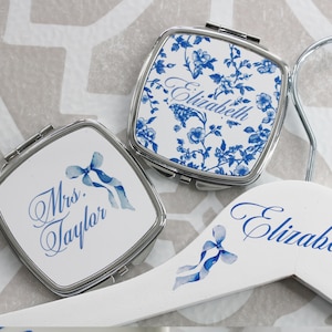 May include: Two compact mirrors with blue and white floral designs. One mirror has the text "Mrs. Taylor" and the other has the text "Elizabeth". A white wooden hanger with the text "Elizabeth" and blue floral accents.
