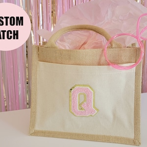 May include: A small, beige burlap tote bag with a white canvas pocket featuring a pink and gold letter Q. The bag has a pink straw sticking out of the top.