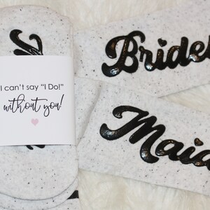 K&ouml;nnte beinhalten: Zwei Paar wei&szlig;e Socken mit schwarzer Beschriftung. Ein Paar sagt "Brides" und das andere sagt "Maid". Ein wei&szlig;es Etikett ist an einem Paar Socken befestigt, auf dem steht "Ich kann nicht "Ich tue es!" sagen, ohne dich!"