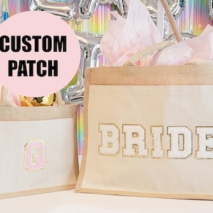 May include: Two burlap tote bags with custom patches. The larger bag has the word "BRIDE" in white glitter letters on a beige background. The smaller bag has a pink letter "Q" on a beige background.